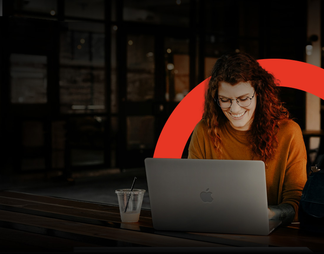 Una mujer sonriente con gafas trabaja en un MacBook Pro en una cafetería, destacada por un arco rojo de diseño que simboliza las soluciones de renting tecnológico.